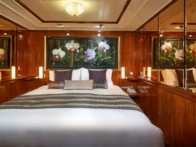 Eine elegante Suite an Bord der Norwegian Gem mit einem großen Doppelbett, umgeben von glänzenden Holzpaneelen und Spiegelflächen. Über dem Bett hängt ein großes Gemälde mit weißen Orchideen, flankiert von Nachttischlampen und einem Telefon.