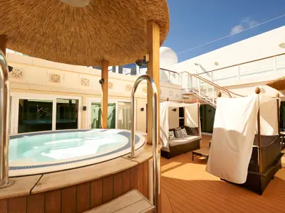 An Bord der Norwegian Gem: Privater Suiten-Außenbereich mit großem Whirlpool und modernen Korbliegen unter Stoffdächern auf sonnigem Holzdeck. Eine Treppe ist ebenfalls sichtbar.