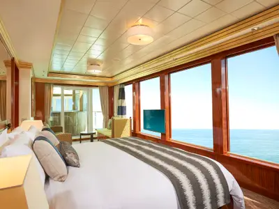 Eine helle Suite an Bord der Norwegian Gem. Im Vordergrund ein großes Bett, dahinter bodentiefe Fenster mit weitem Meerblick. Ein TV ist am Fenster montiert.