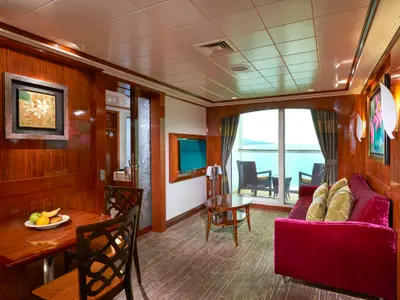 Wohnbereich einer luxuriösen Suite an Bord der Norwegian Gem. Sichtbar sind ein Esstisch mit Früchten, ein rotes Samtsofa mit Kissen, ein Fernseher und ein Balkon mit Meerblick.