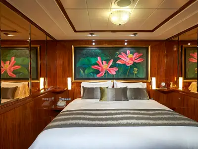 Master-Kabine an Bord der Norwegian Gem: Ein luxuriöses Doppelbett, darüber ein großes Gemälde mit roten Lotusblüten. Dunkle Holzwände und Spiegel sorgen für ein elegantes Ambiente.