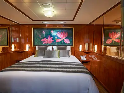 Eine elegante Suite an Bord der Norwegian Gem mit einem grossen Doppelbett, bequemen Kissen und einer gemusterten Tagesdecke. Über dem Bett hängt ein grosses Wandbild mit rosa Seerosen. Die Wände sind mit edlem Holz verkleidet. Rechts und links vom Bett befinden sich Nachttische mit Lampen und Telefonen. Ein langer Spiegel reflektiert den Raum und eine weitere Seerose an der rechten Wand. Vorhänge flankieren den Raum.