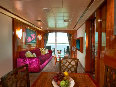 Luxuriöse Suite an Bord der Norwegian Gem mit Wohnbereich, rotem Sofa, Holztäfelung und Balkon mit Meerblick. Im Vordergrund ein Esstisch mit Obst.