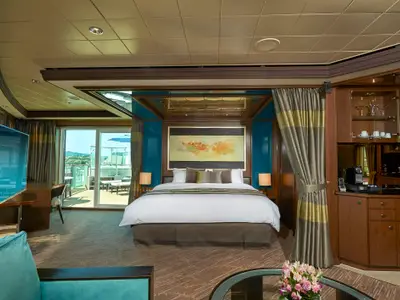 Eine luxuriöse Owners Suite an Bord der Norwegian Gem mit großem Doppelbett, Schreibtisch, Fernseher, Minibar und Zugang zu einem privaten Balkon mit Meerblick.