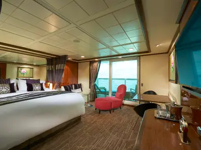 Elegante Suite an Bord der Norwegian Gem mit großem Bett, rotem Sessel, Schreibtisch und privatem Balkon. Vom Balkon aus hat man einen weiten Meerblick. Die luxuriöse Einrichtung wirkt einladend.