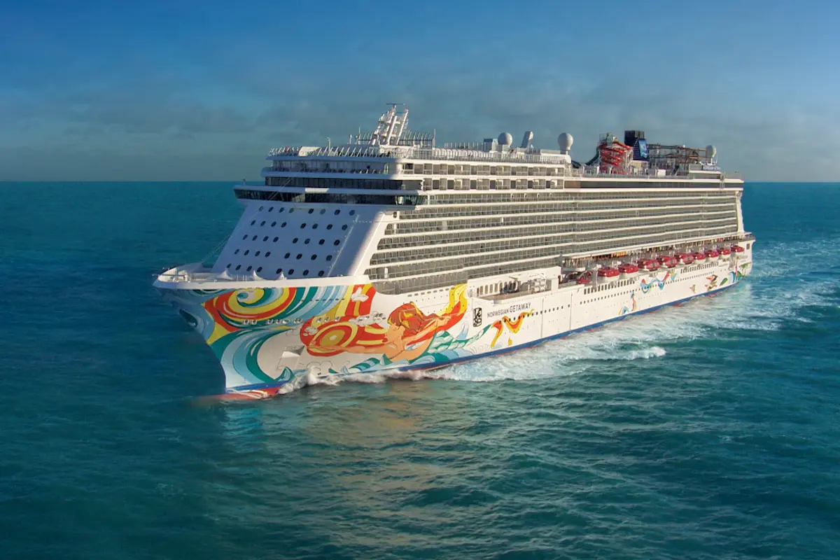 Die Norwegian Getaway gleitet durch das tuerkisfarbene Meer, das detaillierte Bug mit Meerjungfrau-Kunst ist sichtbar. Im Hintergrund der blaue Himmel mit wenigen Wolken.