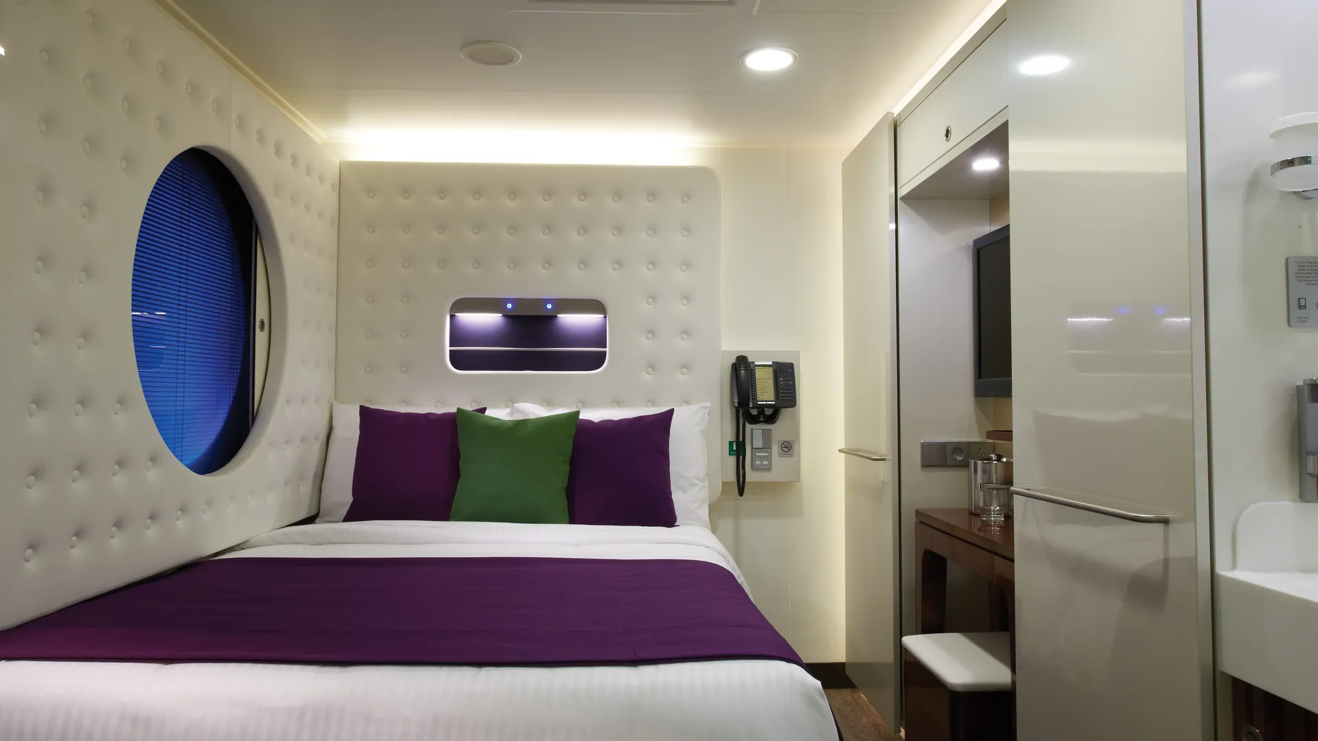 Moderne Studio-Einzelkabine an Bord der Norwegian Getaway. Bett mit lila Decke und Kissen, rundes Fenster mit Jalousie, Schreibtisch, TV und Waschbecken.