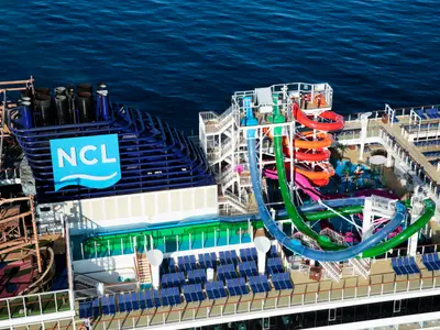 Eine dynamische Vogelperspektive zeigt die bunten Wasserrutschen, Sportdecks und den Schornstein mit NCL-Logo an Bord der Norwegian Getaway auf blauem Meer.