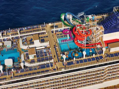 Vogelperspektive auf das sonnige Oberdeck der Norwegian Getaway. Zu sehen sind mehrere Pools, bunte Wasserrutschen, eine Joggingbahn und viele Sonnenliegen mit Gästen am Meer. Ein lebhaftes Deck mit viel Unterhaltung.