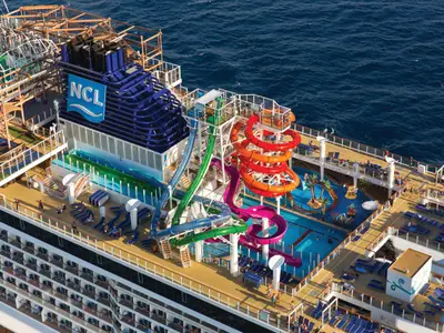 Luftaufnahme des belebten Oberdecks der Norwegian Getaway. Wasserrutschen, Pools und ein Klettergarten sind zu sehen. Passagiere genießen die Norwegian Getaway an Bord.