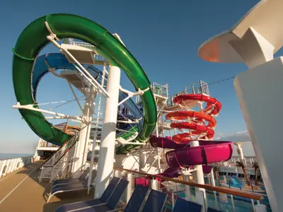 Der AquaPark der Norwegian Getaway: grüne, blaue, rote Spiral- und lilafarbene Wasserrutschen unter blauem Himmel. Meer und Sonnendeck mit Liegestühlen sind sichtbar.