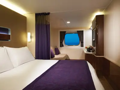 Eine moderne Aussenkabine an Bord der Norwegian Getaway. Ein weiss bezogenes Doppelbett mit violetter Decke dominiert den Vordergrund. Rechts daneben ist eine Wand mit einer TV-Einheit und einem Schreibtisch zu sehen. Am Fenster in der Mitte des Bildes befindet sich eine gemütliche Sitzbank. Durch das runde Fenster ist das blaue Meer sichtbar.