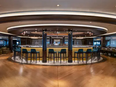 Eine elegant gestaltete Bar namens 'Prime Meridian' an Bord der Norwegian Getaway. Die runde Bar hat goldene Akzente, eine Marmorplatte und ist umgeben von tealfarbenen Barhockern auf einem hellen Holzboden. Dahinter sind diverse Spirituosenflaschen zu sehen, und der Raum ist warm beleuchtet.