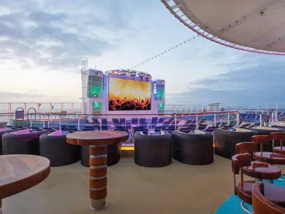 Das Spice H2O Deck der Norwegian Getaway bei Abenddaemmerung. Grosser LED-Bildschirm zeigt eine Konzertszene. Auf dem Deck: Lounge-Moebel, Tische, Barhocker. Pool und Meer im Hintergrund sichtbar.