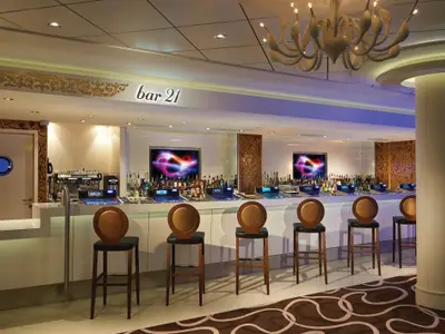 Die Bar 21 an Bord der Norwegian Getaway. Heller Bartresen mit Spirituosen, Bildschirmen, Barhockern. Goldene Ornamente und ein Kronleuchter praegen den eleganten Innenraum.