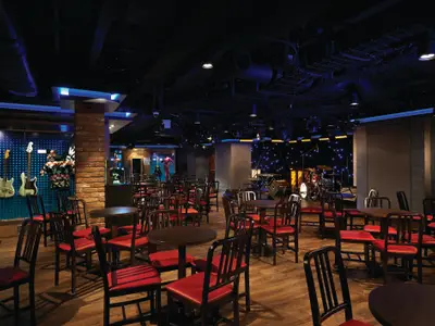 Die GRAMMY Experience Lounge an Bord der Norwegian Getaway. Ein abgedunkelter Raum mit Holzboden, vielen runden Tischen und roten Stühlen. Eine Bühne mit Instrumenten und an der Wand hängende Gitarren.