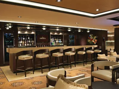 Die elegante The Haven Bar und Lounge an Bord der Norwegian Getaway. Lange Theke, Barhocker, Flaschenregale und bequeme Sessel auf Teppich. Warme Beleuchtung.