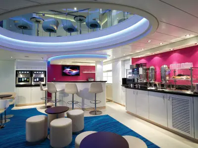 Moderne Lounge an Bord der Norwegian Getaway mit Bar, Automaten und Snackbar. Pinke Wand, blauer Teppich und offene Galerie mit Sitzplätzen prägen den Raum.