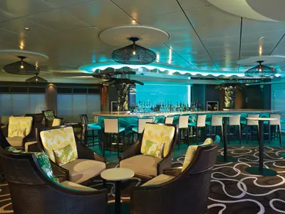 Die Sugarcane Bar an Bord der Norwegian Getaway zeigt eine stilvolle Lounge mit tropischem Dekor, Rattanmöbeln, gemusterten Kissen und einer beleuchteten Bar mit einer großen Auswahl an Getränken.