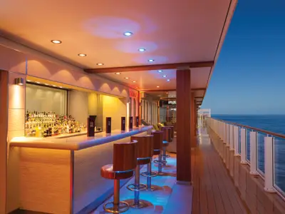 Eine einladende Outdoor-Bar an Bord der Norwegian Getaway bei Abenddämmerung, mit bunten Lichtern und Meerblick. Leere Barhocker und eine große Auswahl an Flaschen.
