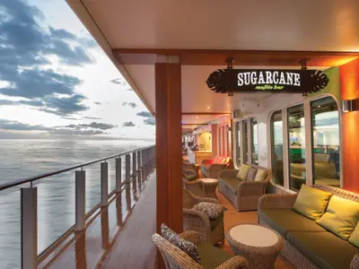 Aussenbereich der Sugarcane Mojito Bar an Bord der Norwegian Getaway mit Rattanmöbeln. Meerblick unter bewölktem Himmel. Beleuchtetes 'Sugarcane' Schild über dem Tresen.