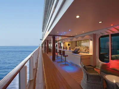 Eine stimmungsvolle Outdoor-Bar und Lounge mit blauer und roter Beleuchtung an Bord der Norwegian Getaway. Rechts sind bequeme Korbsessel, links das weite Meer sichtbar. Der Bereich ist unbesetzt.
