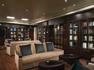 Eine elegante Bibliothek an Bord der Norwegian Getaway mit dunklen Holzregalen voller Buecher und beigen Sofas mit blauen Kissen.