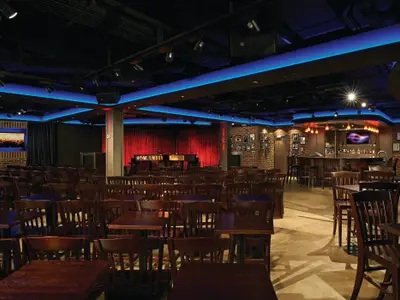 Innenansicht der Howl at the Moon Piano Bar an Bord der Norwegian Getaway. Man sieht die Bühne mit zwei Pianos, davor dunkle Tische und Stühle sowie eine beleuchtete Bar mit Hockern. Blaue LED-Beleuchtung an der Decke sorgt für Stimmung.