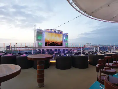 Abend auf der Norwegian Getaway. Große LED-Leinwand zeigt Konzert auf dem Deck. Viele Sitzgelegenheiten und Tische. Meer und bewölkter Himmel im Hintergrund.