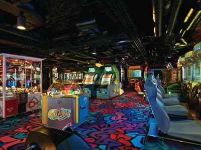 Eine bunte und lebhafte Spielhalle an Bord der Norwegian Getaway. Zahlreiche Arcade-Automaten mit Rennspielen, Greifautomaten und anderen Spielen sind unter einer dunklen Decke mit freiliegenden Rohren angeordnet. Der Teppich hat ein auffälliges, farbenfrohes Muster.
