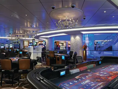 Weitwinkelansicht des eleganten Casinos an Bord der Norwegian Getaway. Im Vordergrund ein Craps-Tisch mit gestapelten Chips. Zahlreiche weitere Spieltische und Reihen von Spielautomaten sind sichtbar. Moderne Kronleuchter und blaue Lichtakzente schmücken den Innenraum. Eine geschwungene Treppe führt in einen tieferliegenden Bereich.