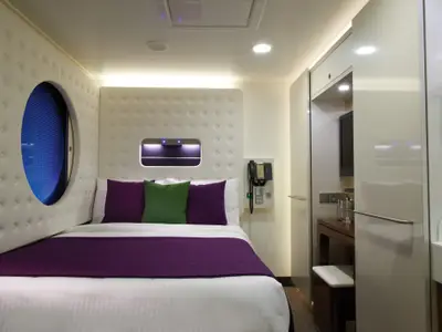 Moderne Studio-Einzelkabine an Bord der Norwegian Getaway. Bett mit lila Decke und Kissen, rundes Fenster mit Jalousie, Schreibtisch, TV und Waschbecken.