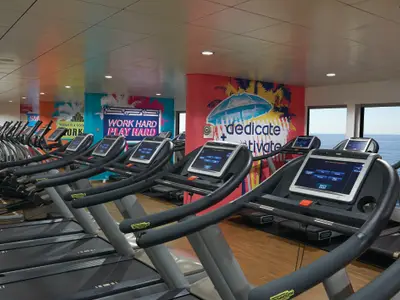 Ein modernes Fitnesscenter an Bord der Norwegian Getaway mit mehreren Reihen von Laufbaendern und Crosstrainern. Grosse Fenster bieten einen weiten Blick auf das offene Meer.