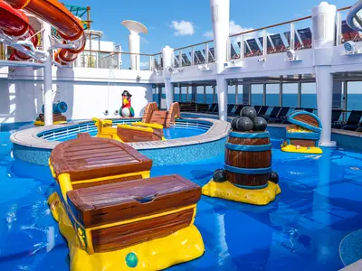 Detailansicht des farbenfrohen Kinder-Wasserparks an Deck der Norwegian Getaway. Piraten-Spielgeräte wie Schatzkisten, Kanonen und ein Papagei umgeben ein Schwimmbecken mit Rutschen. Blauer Boden, im Hintergrund das offene Meer.