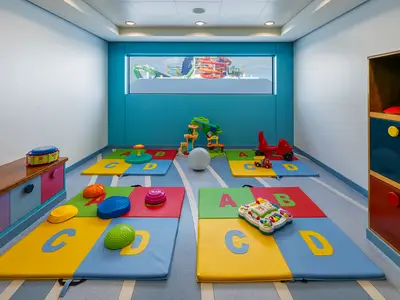 Ansicht eines farbenfrohen Kinderbereichs an Bord der Norwegian Getaway mit vielen Spielmatten, Baukloetzen und Spielzeug. Ein Fenster bietet Blick auf aufregende Wasserrutschen des Schiffs.