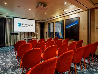 Konferenzraum an Bord der Norwegian Getaway. Rote Stühle blicken auf eine Projektionsfläche mit NCL-Logo. Stehpult und Blumen am Tisch runden das Bild ab.