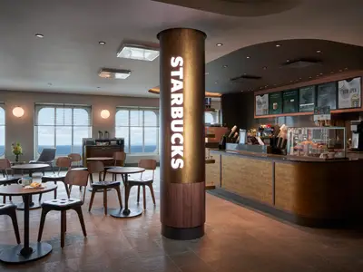 Starbucks-Café an Bord der Norwegian Getaway. Beleuchtetes Logo, eine Theke mit Gebäck und ein Sitzbereich mit Holztischen und Stühlen vor großen Fenstern mit weitem Meerblick.