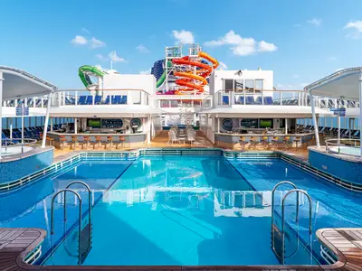 Weitwinkelaufnahme des lebhaften Pooldecks der Norwegian Getaway mit zwei Bars, einem grossen Pool und mehrfarbigen Wasserrutschen unter klarem Himmel.