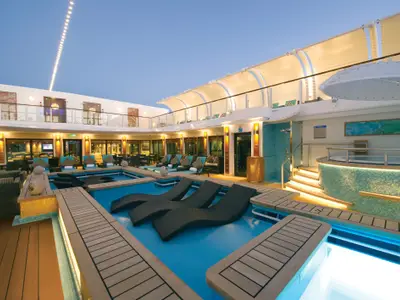 Der luxuriöse Haven Courtyard Poolbereich der Norwegian Getaway bei Abend. Blaue Pools mit Unterwasser-Liegen, Holzböden und ein Whirlpool. Obere Ebene mit Lounge-Sesseln unter Segeldach beleuchtet.