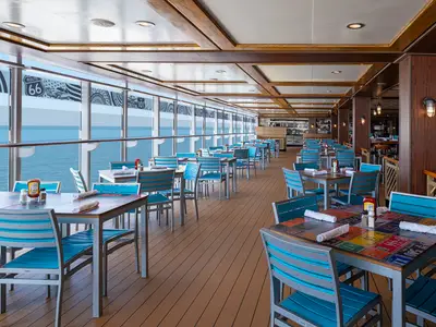 Ein lichtdurchflutetes American Diner-Restaurant an Bord der Norwegian Getaway mit vielen blauen Tischen und Stühlen. Große Fensterfronten bieten einen weiten Blick auf das offene Meer.