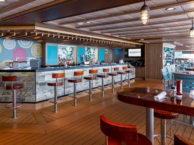Moderne Bar-Lounge an Bord der Norwegian Getaway mit langer Theke, Holz-Hockern und Restauranttischen. Stylische Wanddeko und Holz-Stein-Akzente prägen das Design.