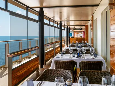 Blick in Cagney's Steakhouse an Bord der Norwegian Getaway. Elegant gedeckte Tische stehen entlang einer Glasfront mit weitem Meerblick. Braune Korbstühle ergänzen die Holzvertäfelung und weiße Tische.