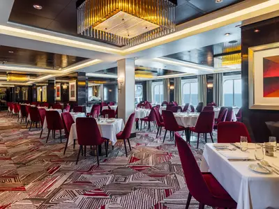 Elegantes Hauptrestaurant Savor an Bord der Norwegian Getaway. Rote Samtsessel an weiss gedeckten Tischen. Grosse Fenster zeigen das Meer. Moderne Einrichtung mit Deckenleuchten.