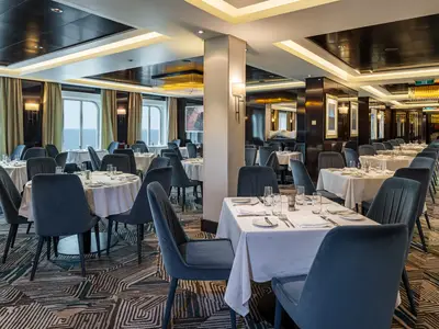 Innenansicht des Restaurants Taste an Bord der Norwegian Getaway. Gedeckte Tische mit blauen Stühlen vor großen Fenstern mit Meerblick. Elegantes Ambiente.