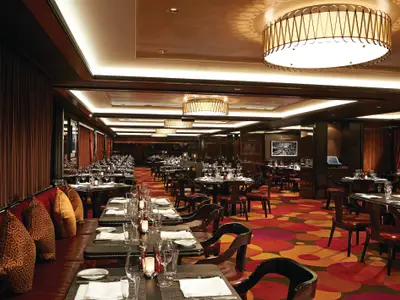 Ein elegantes Spezialitätenrestaurant an Bord der Norwegian Getaway mit vielen gedeckten Tischen. Dunkle Holzelemente, rote Polster und ein gemusterter Teppich dominieren den Raum. Große Leuchter hängen von der Decke.
