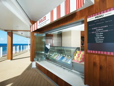 Ein offener Gelato-Stand namens 'Dolce Gelato' an Bord der Norwegian Getaway mit einer Auswahl an Gelato-Sorten in der Auslage und einem Menübrett. Im Hintergrund ist das offene Deck und das Meer unter blauem Himmel zu sehen.