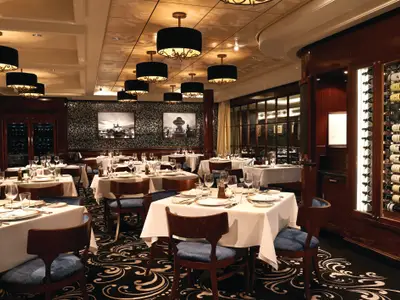 Blick in das edle Spezialitätenrestaurant Le Bistro an Bord der Norwegian Getaway, mit vielen weiß gedeckten Tischen, blauen Stühlen, dunklen Holzelementen und einer Weinwand.