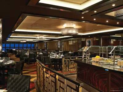 Blick in das elegante Buffetrestaurant Moderno an Bord der Norwegian Getaway. Viele gedeckte Tische und eine Buffettheke mit Speisen füllen den Raum. Meerblickfenster im Hintergrund.