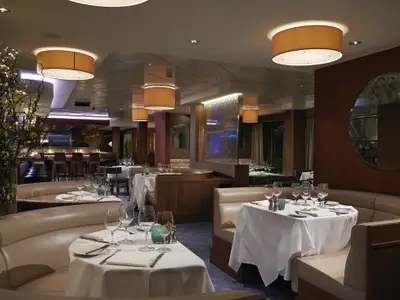 Elegantes Restaurant Ocean Blue an Bord der Norwegian Getaway. Weiss gedeckte Tische, beigefarbene Ledersitzbänke und dunkles Holz. Warme Deckenleuchten und eine Bar schaffen gehobenes Ambiente.