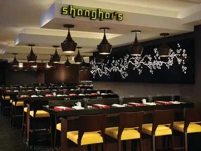 Blick in das Shanghais Spezialitätenrestaurant an Bord der Norwegian Getaway. Dunkle Holzmöbel, gelbe Stühle und eine beleuchtete Kirschblütenwandkunst prägen den Raum.